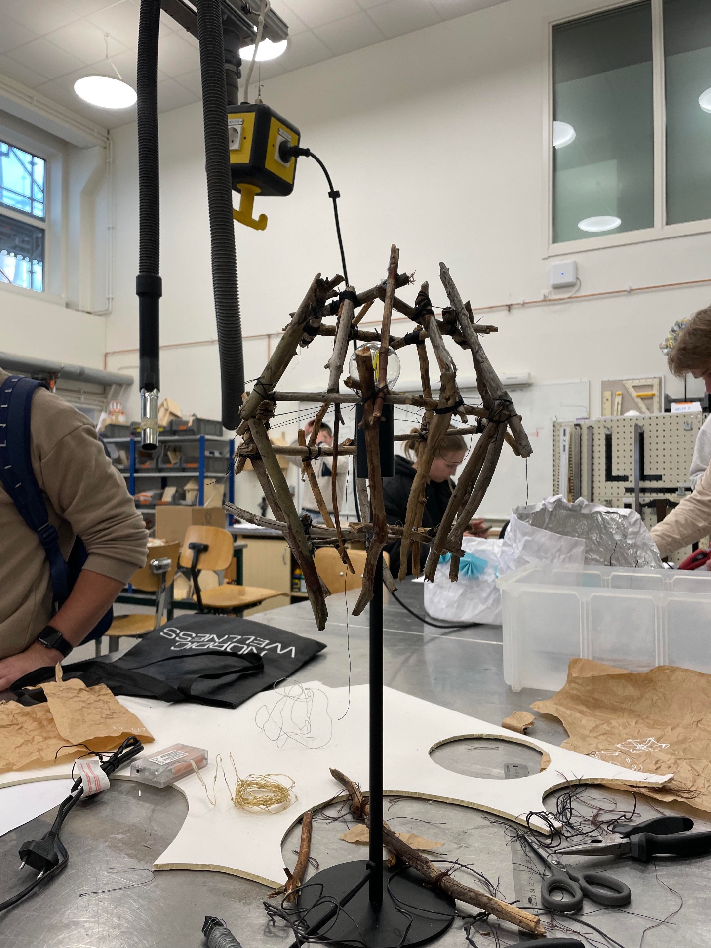 Unfinished branch lamp structure on a stand in a workshop, with materials and tools on the table around it.