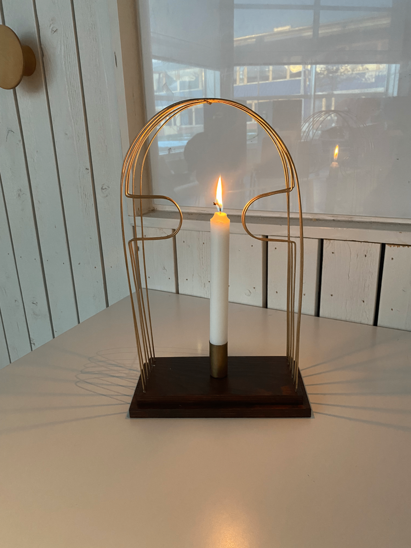 Front view of the finished candle holder with a lit candle at its centre, surrounded by a symmetrical arched metal frame on a stepped dark wooden base.