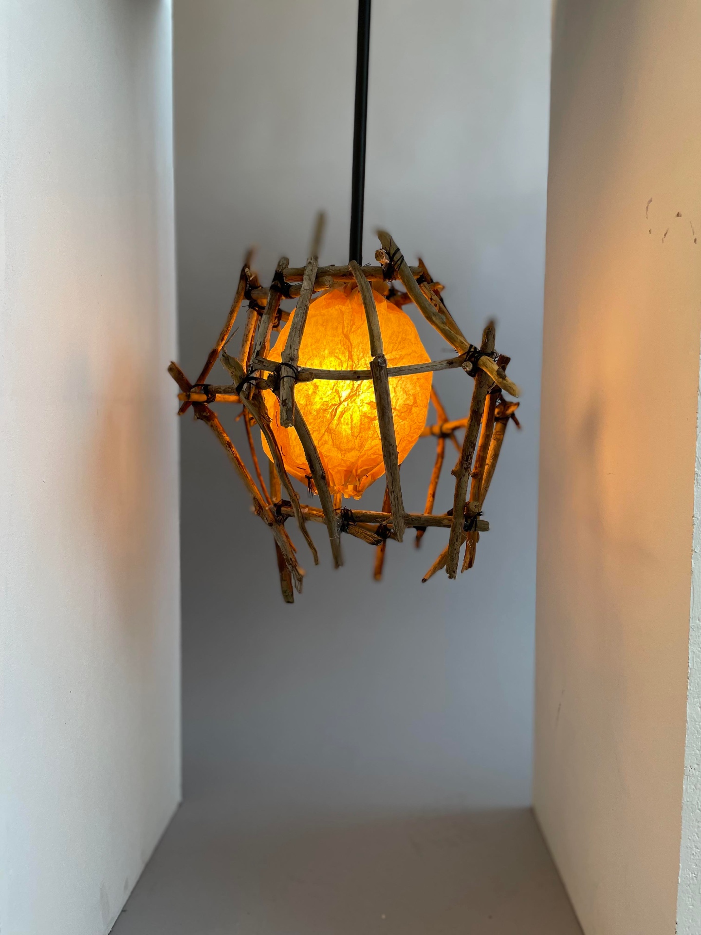 Hanging lamp made from irregular branches tied with string, with a warm paper light element glowing inside, photographed in a narrow white space.
