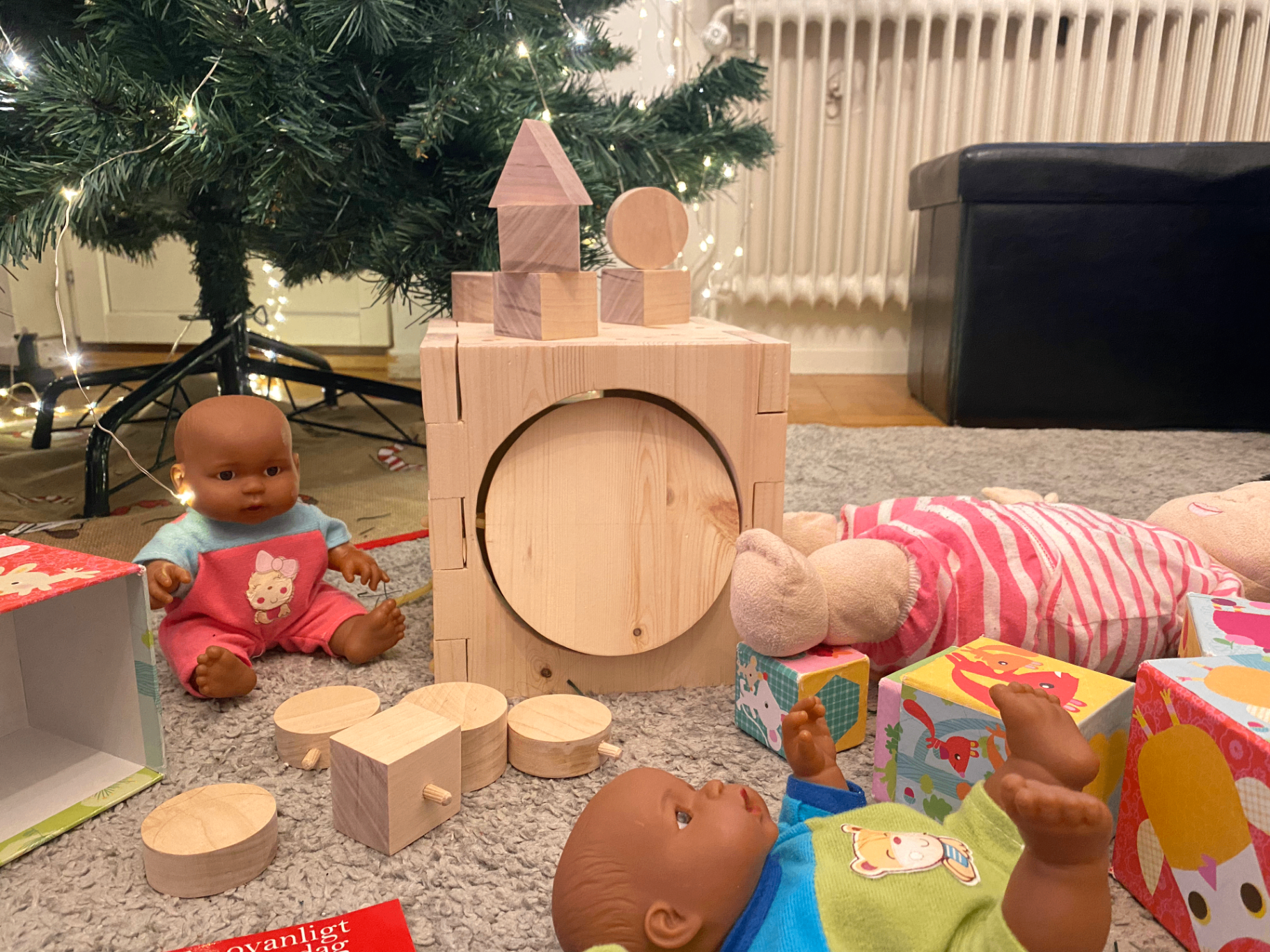 Wooden activity box in a living room with two dolls and colourful toy boxes around it, near a lit Christmas tree.