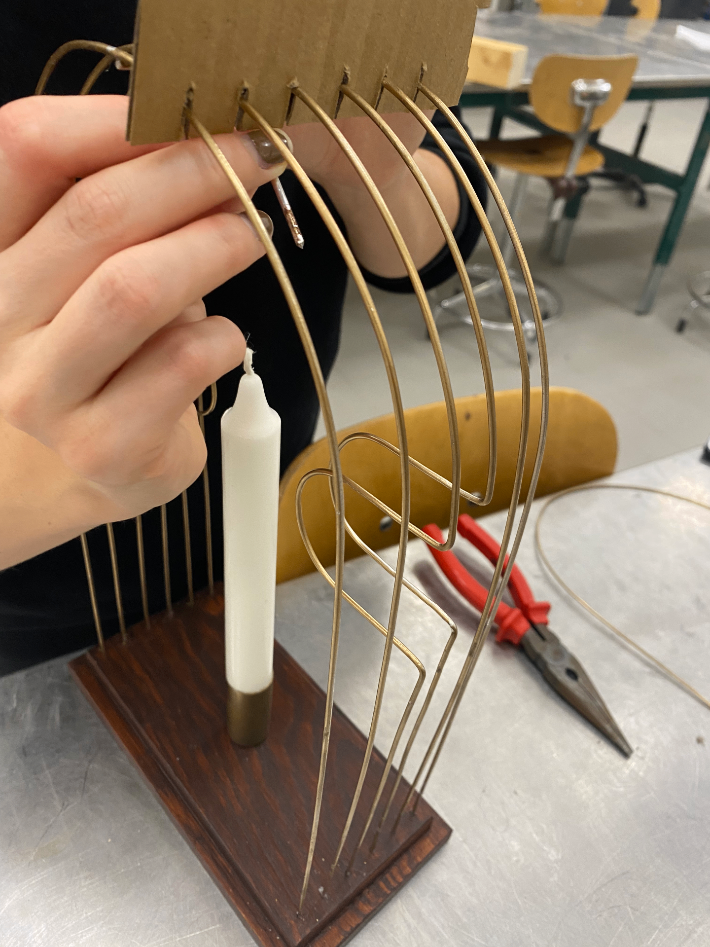 Hand holding a cardboard template above the wooden base while curved metal rods are positioned around a white candle in the workshop.